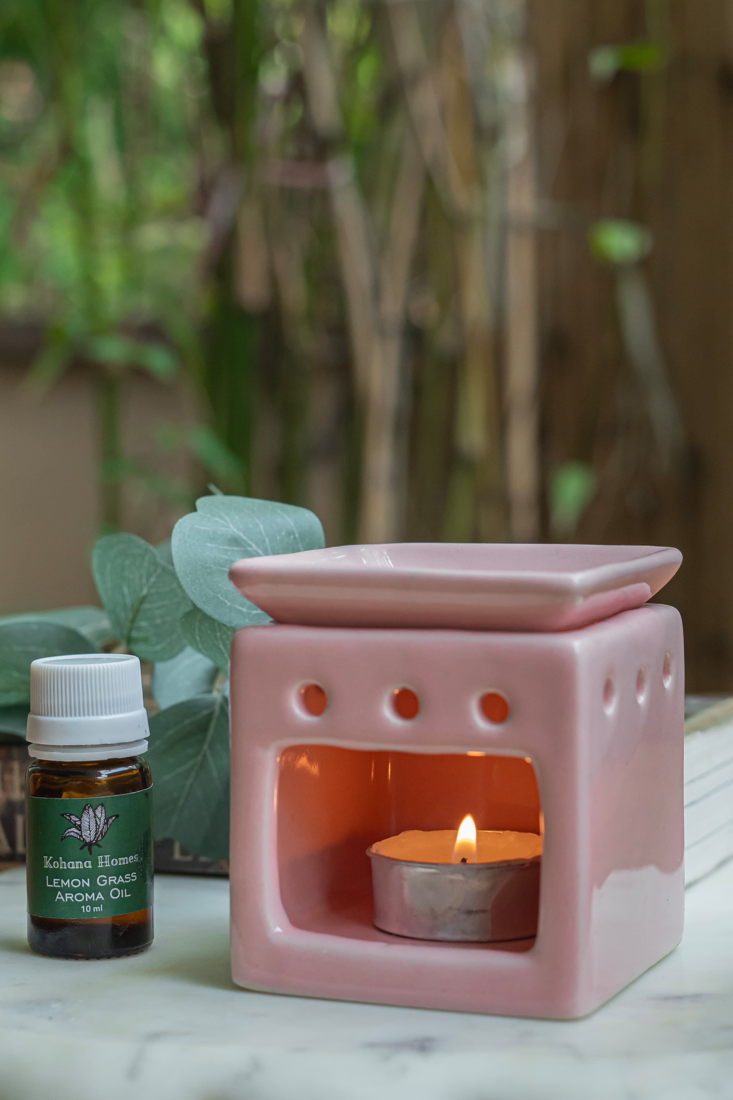 Pink ceramic oil burner with a lit candle and a bottle of essential oil on a marble surface.