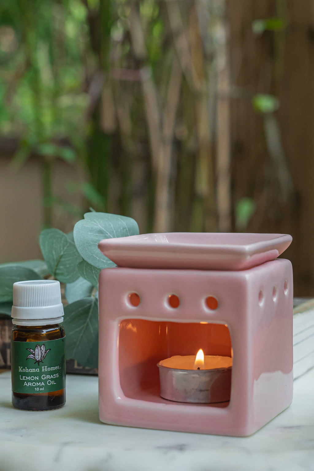 Pink ceramic oil burner with a lit candle and a bottle of essential oil on a marble surface.
