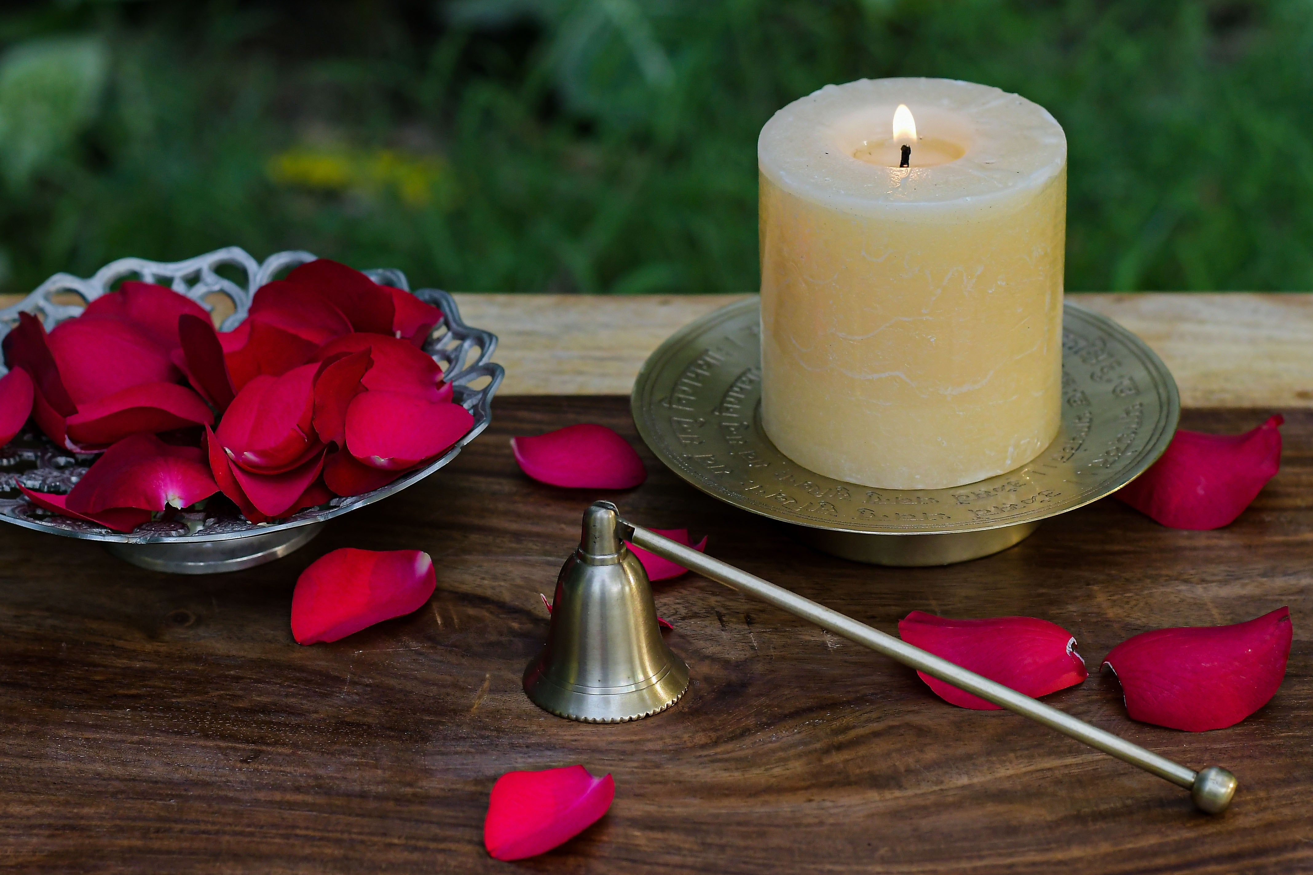 Candle on a metal stand with red rose petals and a snuffer on a wooden surface.