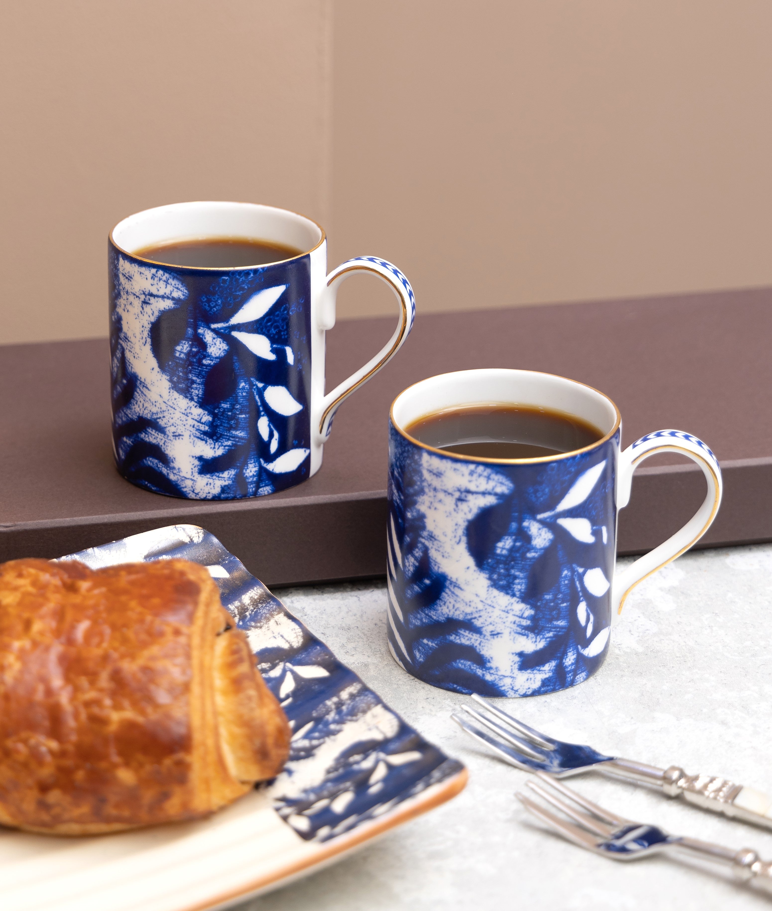 Two blue and white patterned mugs with coffee and a pastry on a tray.