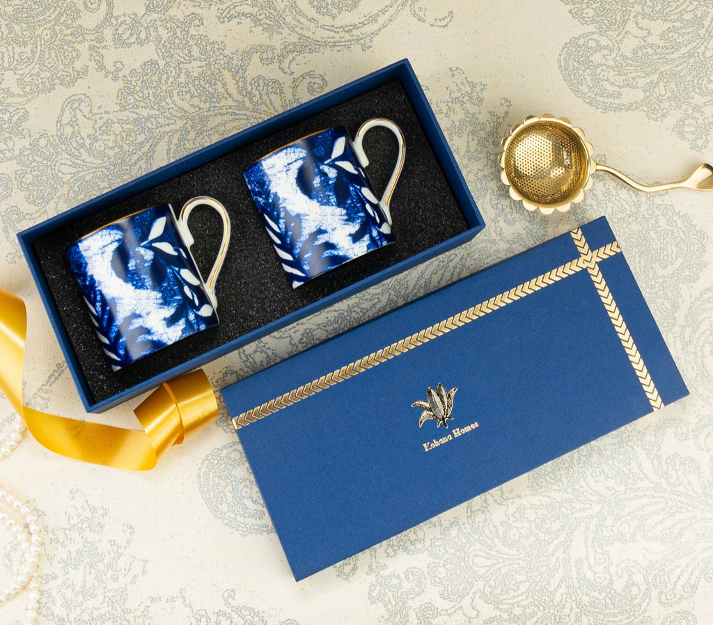 Two blue ceramic mugs in a decorative box with a gold ribbon on a patterned surface.