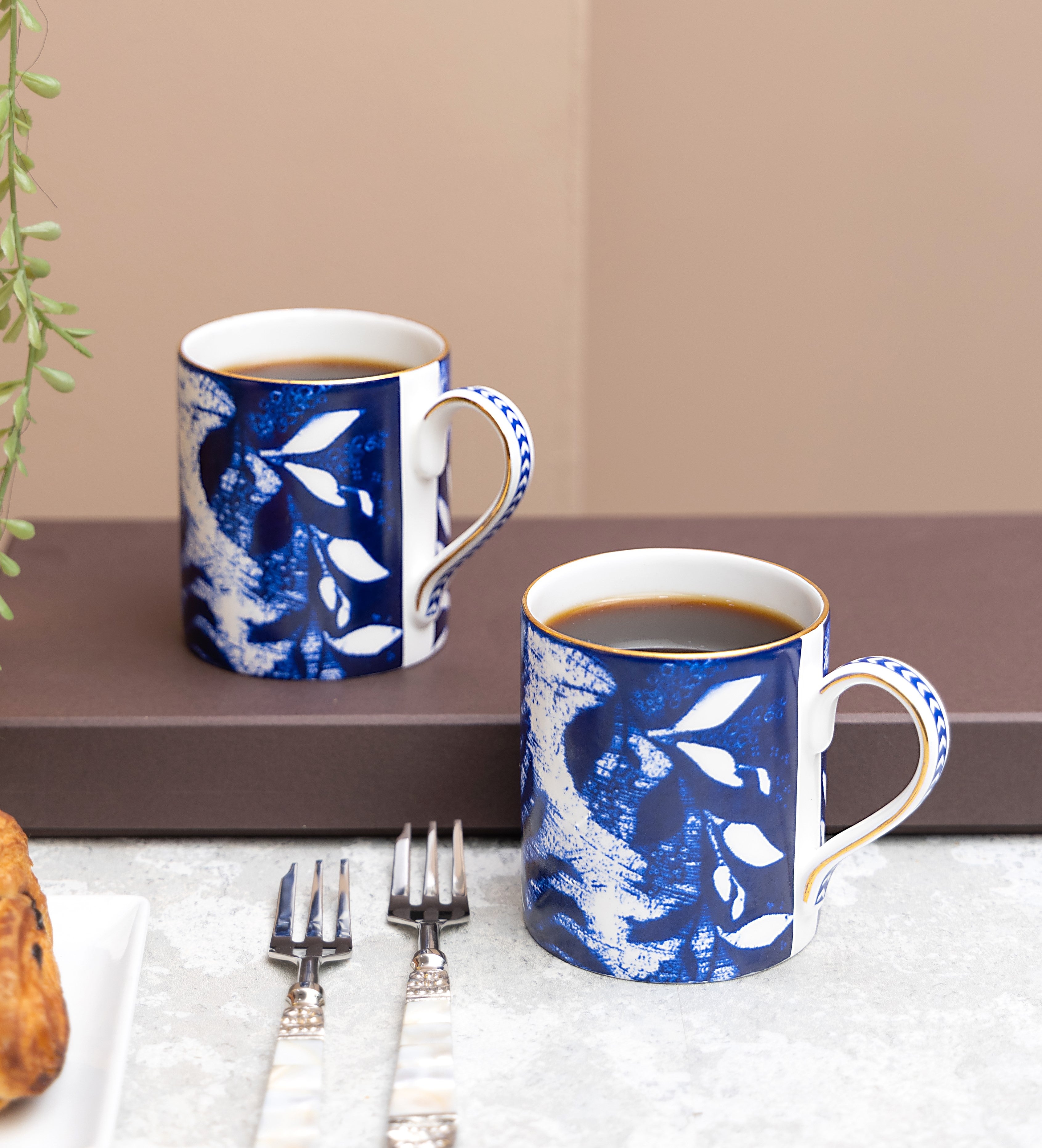 Two blue and white textile print mugs with coffee on a table with cutlery and a plant.