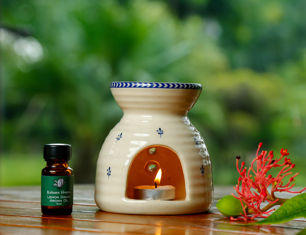 Ceramic oil burner with a lit candle next to a bottle of essential oil on a wooden surface with a blurred green background.