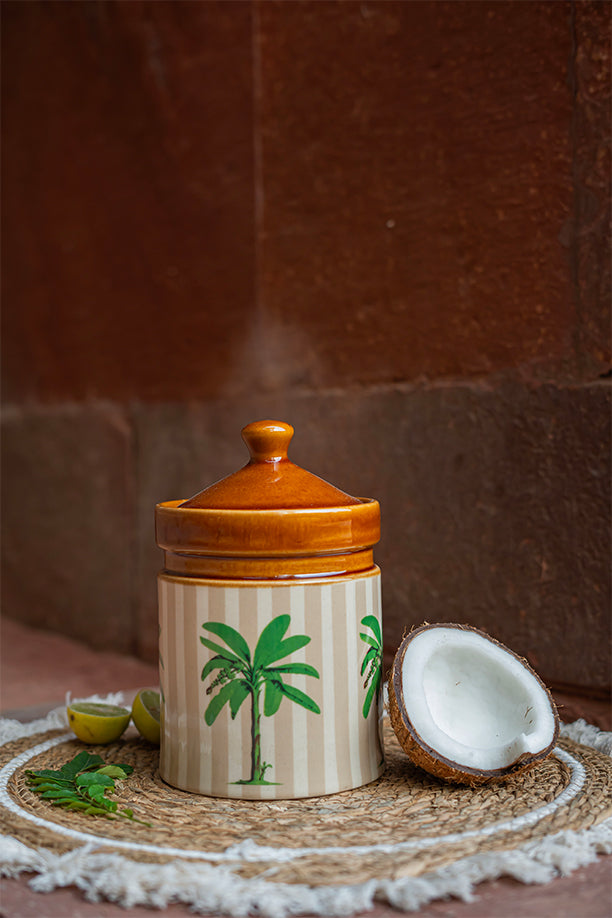 Ceramic jar with a lid on a woven mat with a coconut and leaves on a brown background
