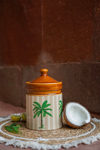 Ceramic jar with a lid on a woven mat with a coconut and leaves on a brown background